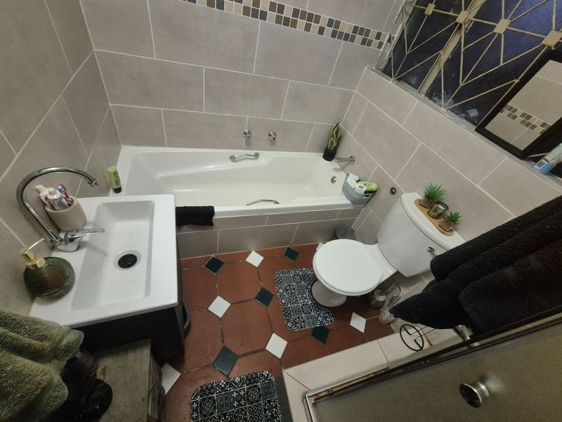 A compact bathroom featuring a built-in bathtub, vanity basin, and toilet with neutral wall tiling and patterned floor tiles.