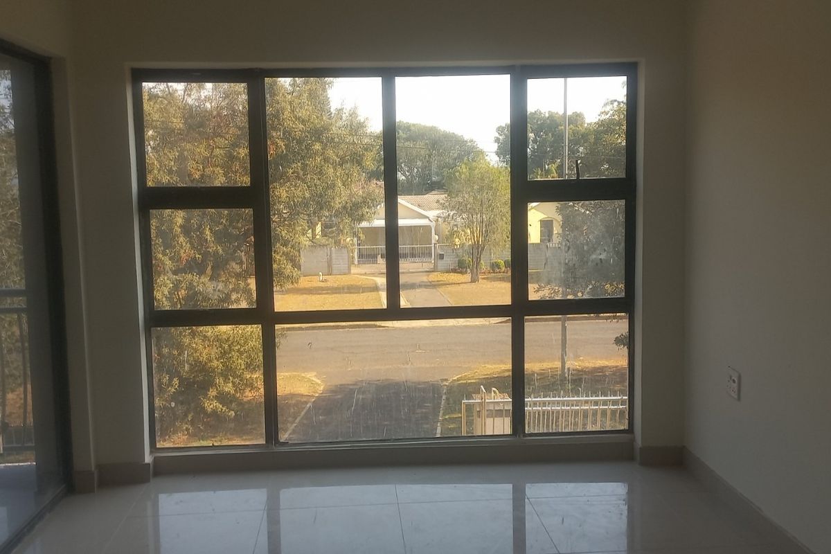 An empty living area featuring large aluminium-framed windows providing natural light and a view of the street, with neutral tiled flooring throughout.