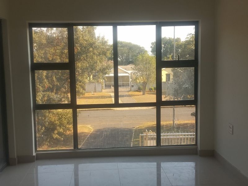 An empty living area featuring large aluminium-framed windows providing natural light and a view of the street, with neutral tiled flooring throughout.