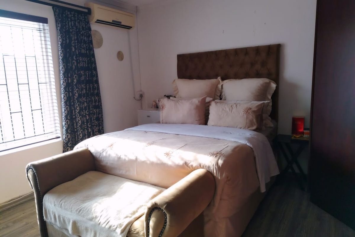 A functional bedroom featuring laminate flooring, a wall-mounted air conditioning unit, and security bars on the window.