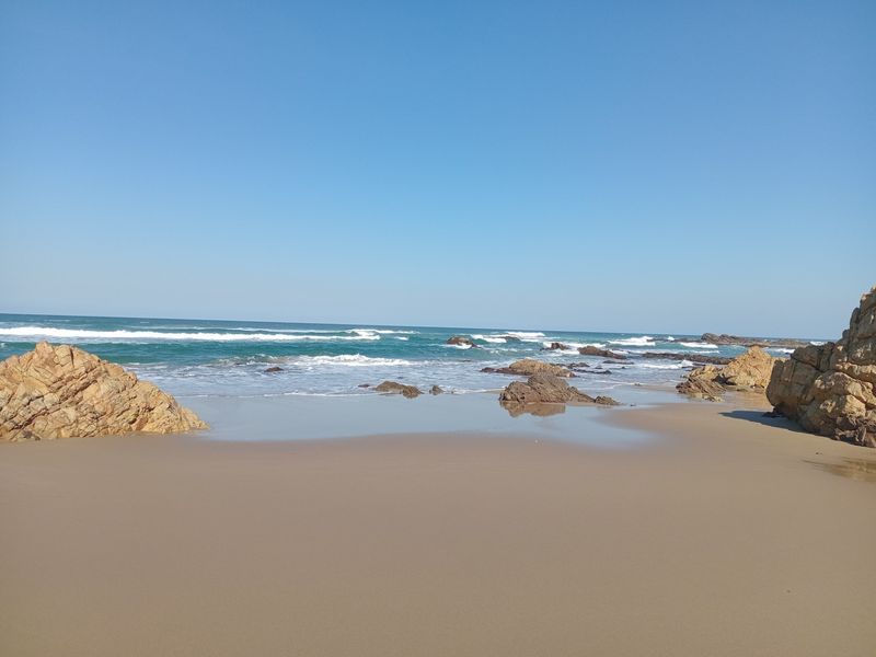 A scenic view of a sandy beach with natural rock formations and ocean waves under a clear blue sky.