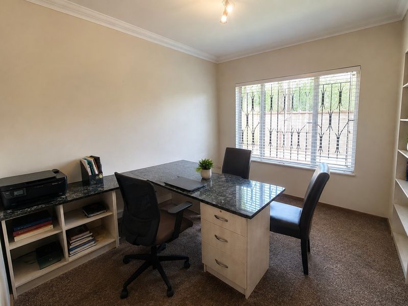 A functional home office featuring built-in desk cabinetry, integrated shelving, carpeted flooring, and a window fitted with security bars and venetian blinds.