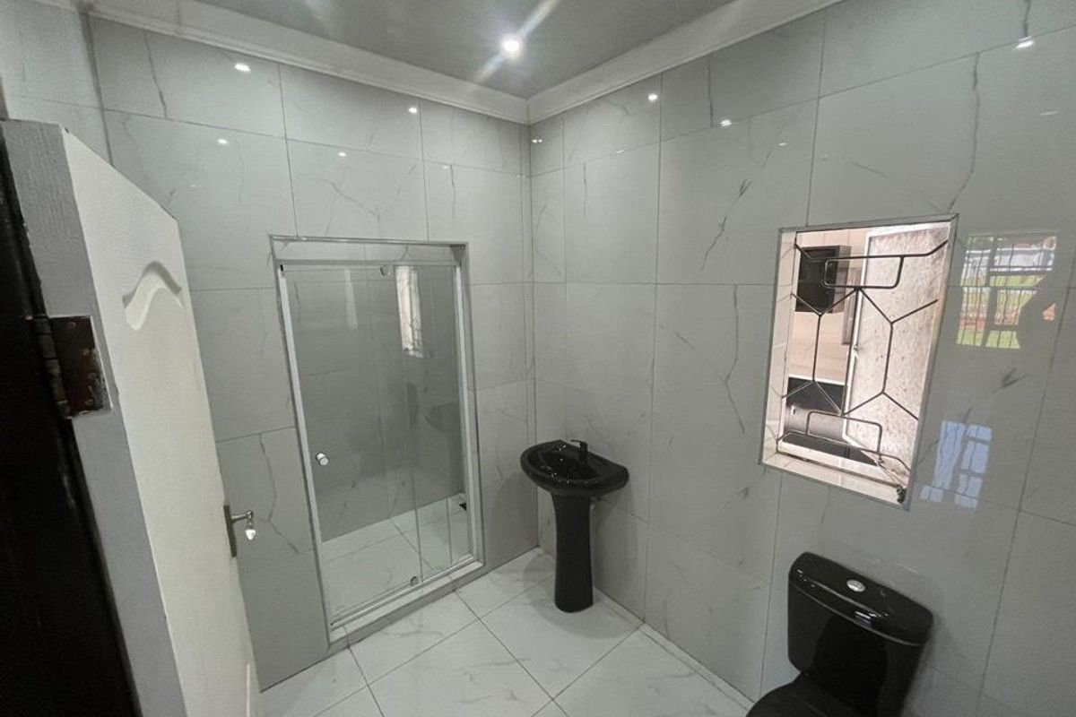 A modern bathroom featuring white marble-look wall and floor tiles, a black pedestal basin, a black toilet, and a glass shower enclosure.