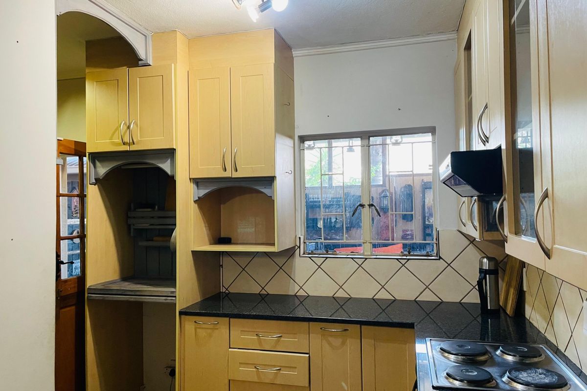 A functional kitchen featuring light wood cabinetry, dark granite countertops, a built-in electric hob, and a tiled backsplash with security-barred window.