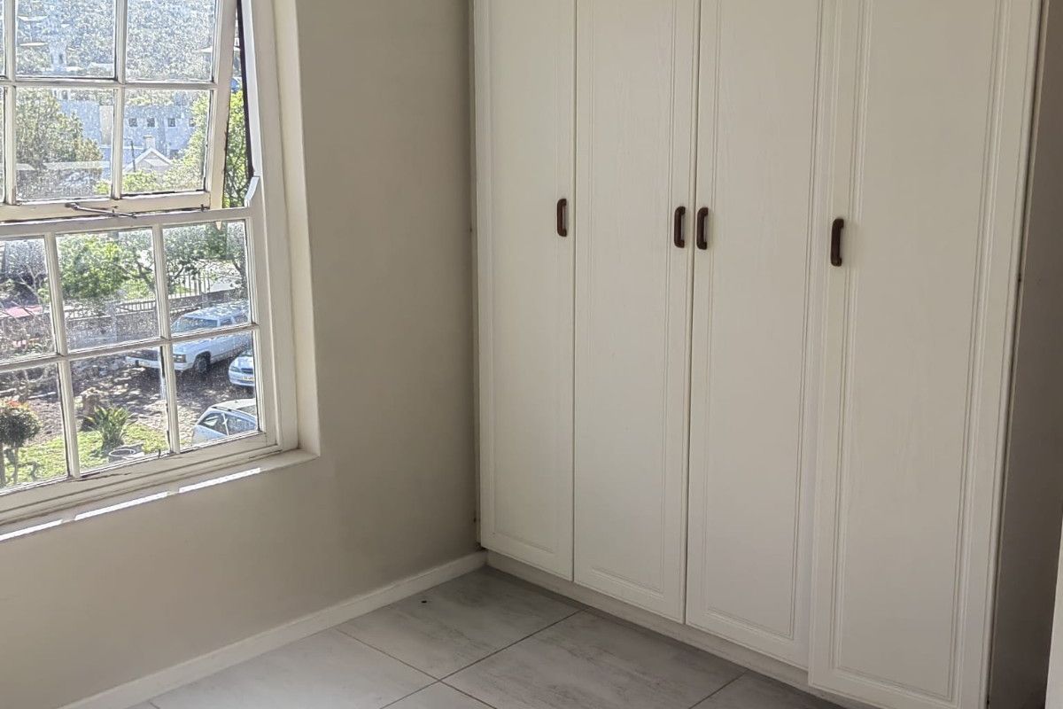 A bright, empty bedroom featuring neutral tiled flooring, built-in white cupboards, and a sash window with natural light.