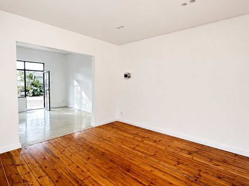 A bright, vacant living room featuring polished hardwood flooring, recessed lighting, and a seamless transition into an adjacent sunlit space.