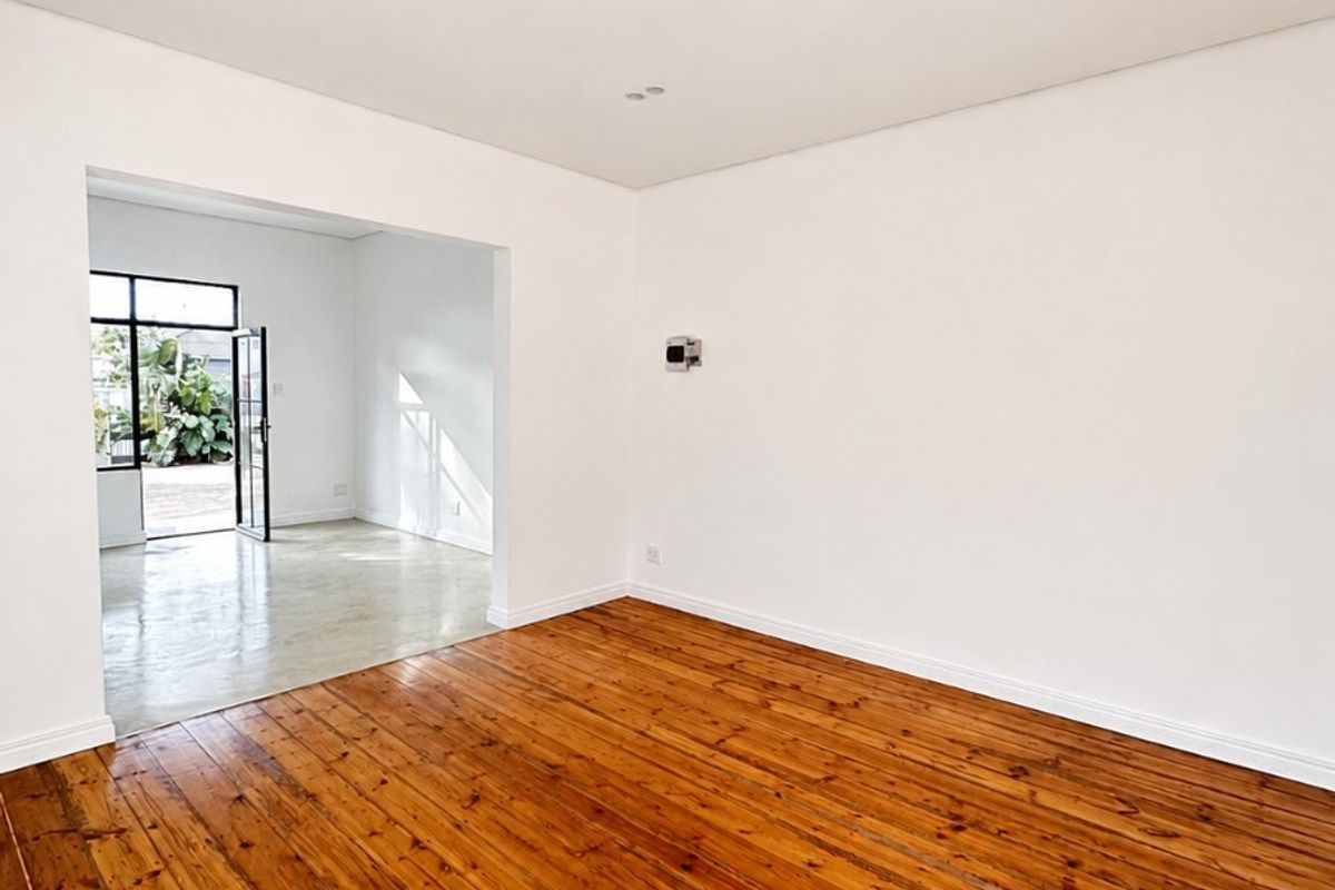 A bright, vacant living room featuring polished hardwood flooring, recessed lighting, and a seamless transition into an adjacent sunlit space.