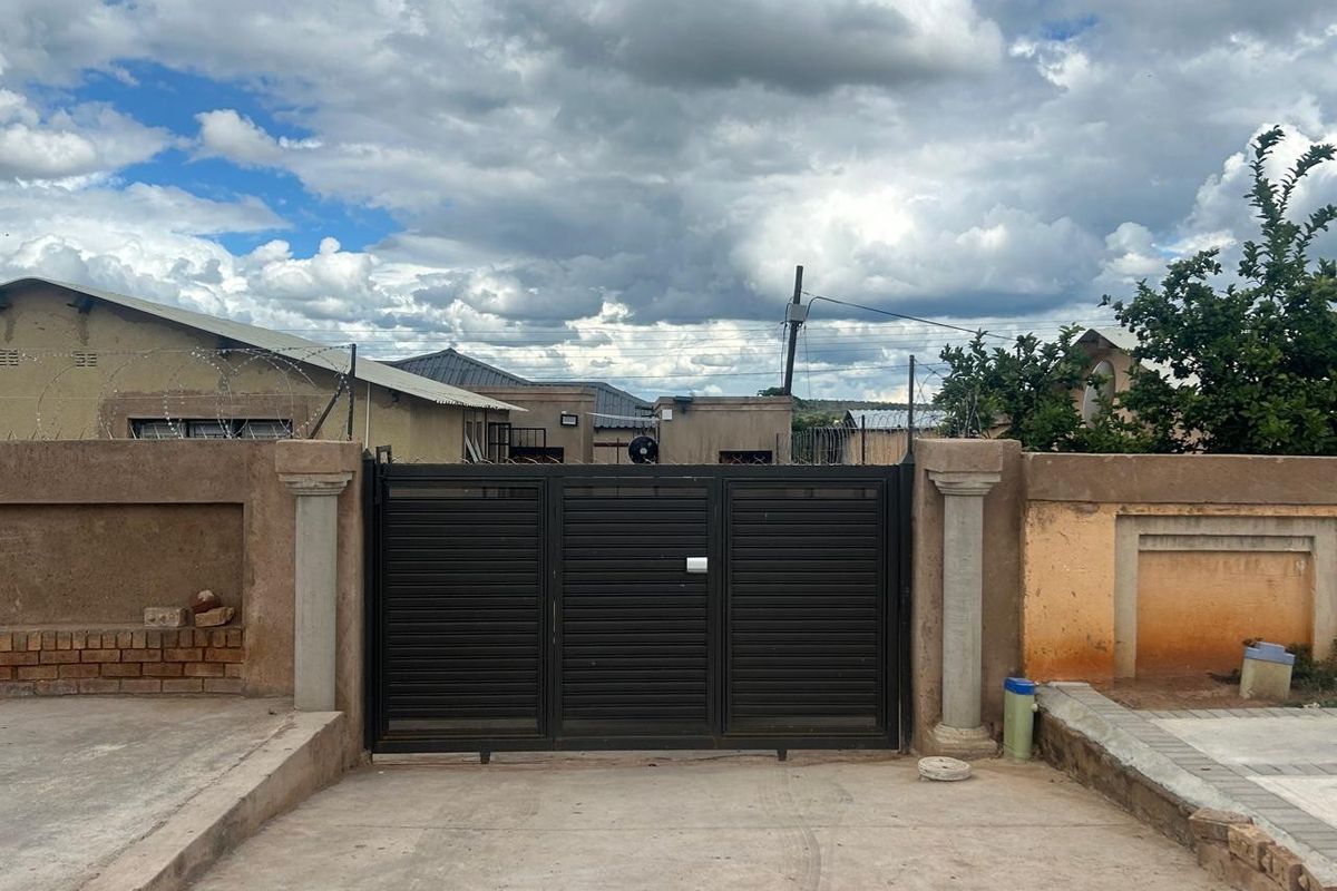 Front view of the property featuring a secure metal driveway gate, perimeter wall with razor wire, and a concrete driveway.
