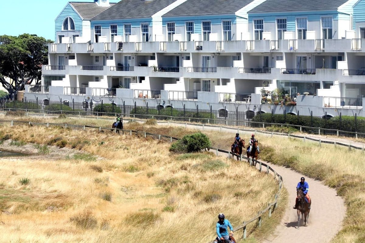 Rear elevation of a multi-story apartment complex featuring private balconies, overlooking a natural landscape with a dirt path.