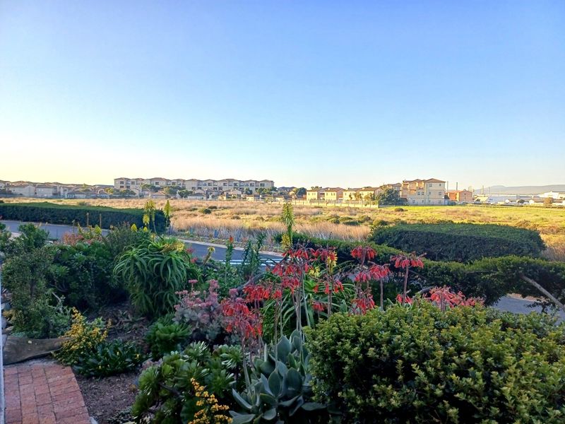 A scenic view overlooking a landscaped garden with native plants, leading to an open field and residential apartment complex in the distance.