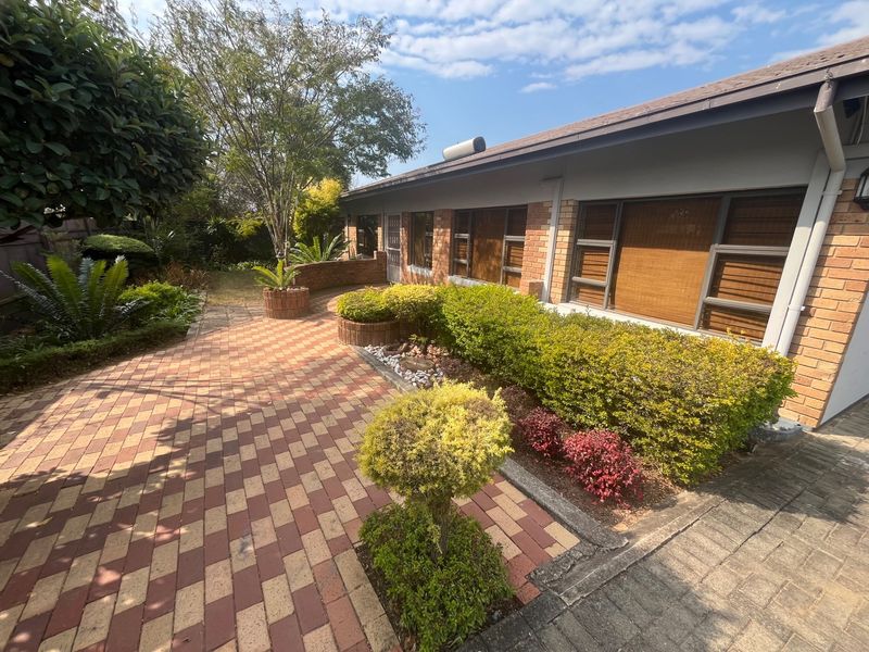 A well-maintained rear exterior view featuring a brick facade, paved patio area, and established garden landscaping with aluminium window frames.