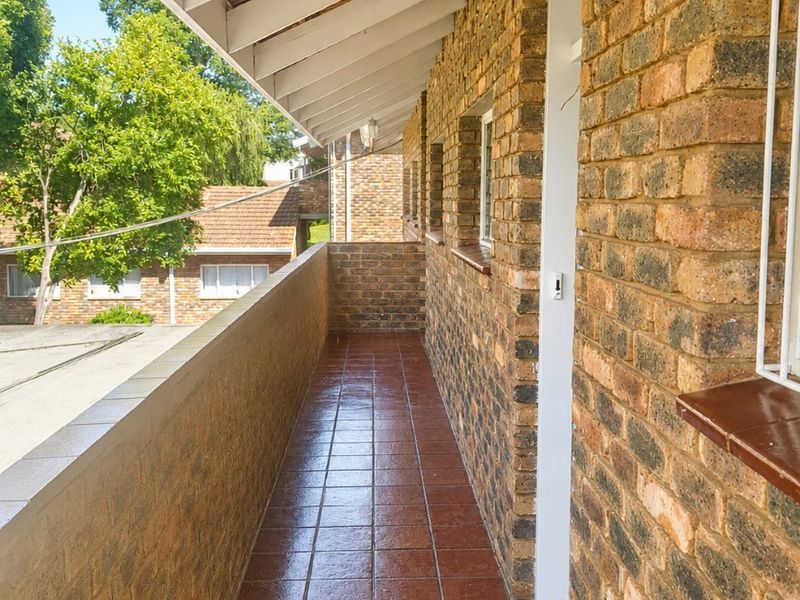 A covered balcony featuring facebrick walls, tiled flooring, and exposed timber rafters, providing an outdoor space with a view of the surrounding complex.