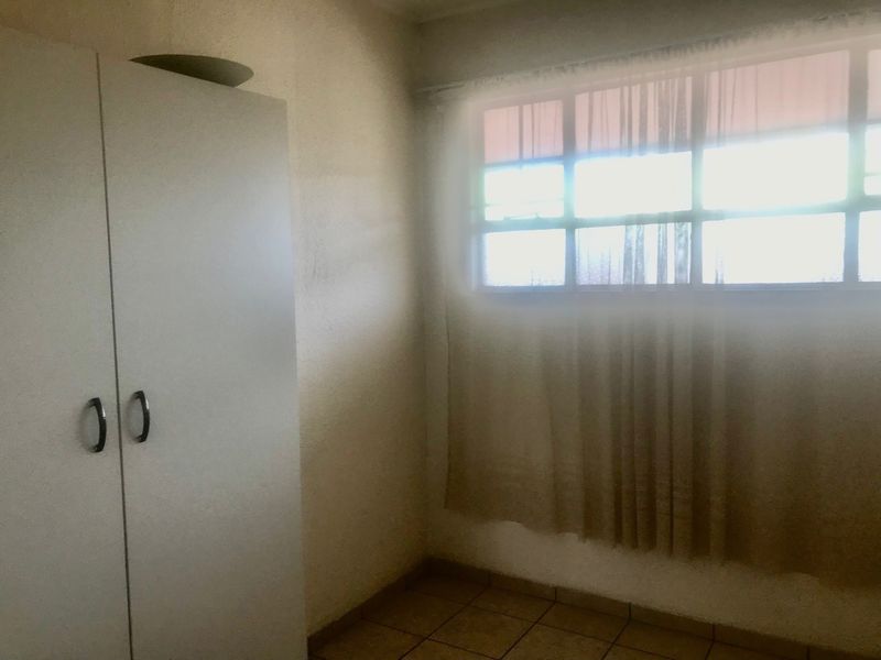 A simple, vacant bedroom featuring tiled flooring and a built-in wardrobe, illuminated by natural light from a large window.