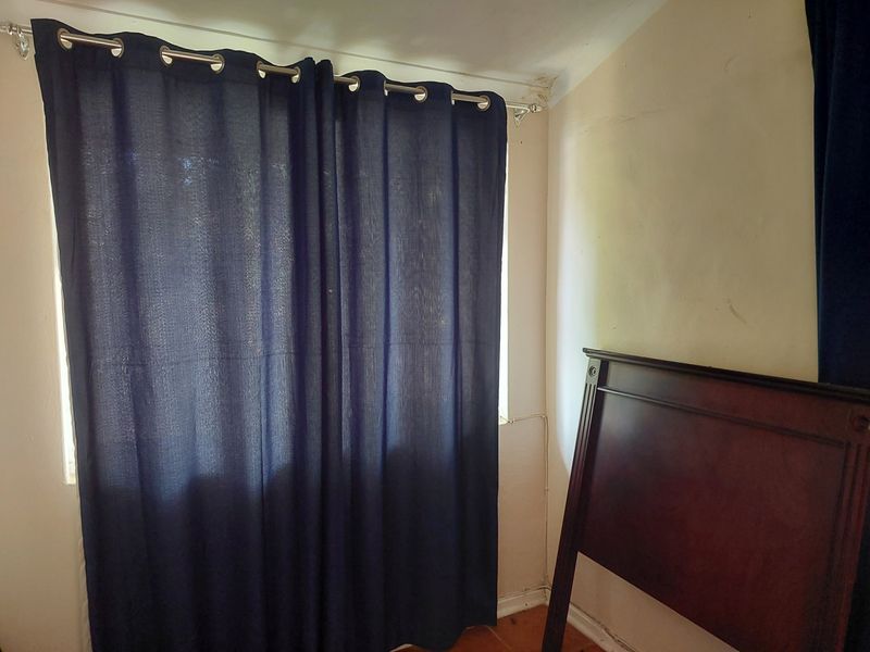 A bedroom corner featuring a window with a curtain rod, showing visible signs of ceiling water damage and wall wear.