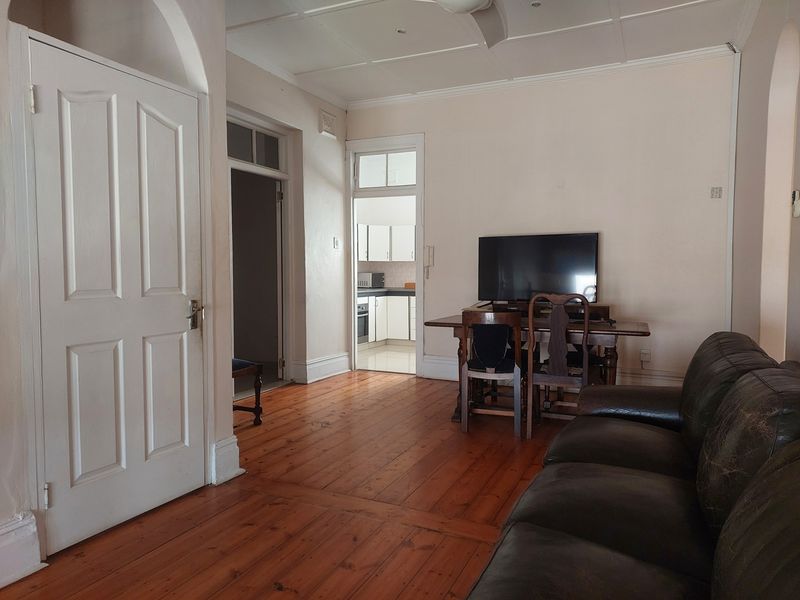 A living room featuring original timber flooring, high ceilings with panelled detailing, and an arched doorway leading to an adjacent kitchen area.