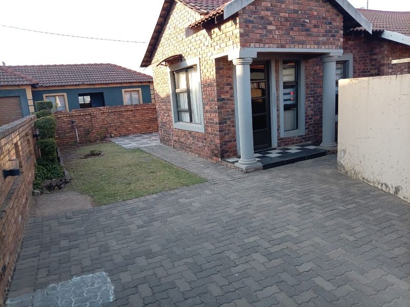 A rear exterior view of a face-brick house featuring a paved driveway, small lawn, and covered entrance with concrete columns.