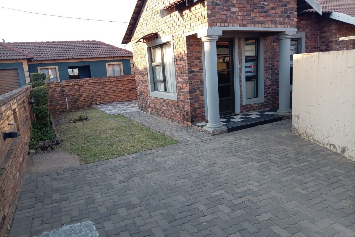 A rear exterior view of a face-brick house featuring a paved driveway, small lawn, and covered entrance with concrete columns.