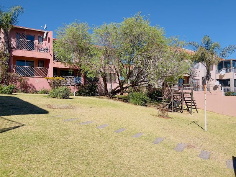 A view of the rear exterior of the townhouse complex, featuring a shared lawn area, mature trees, and balconies with security bars.
