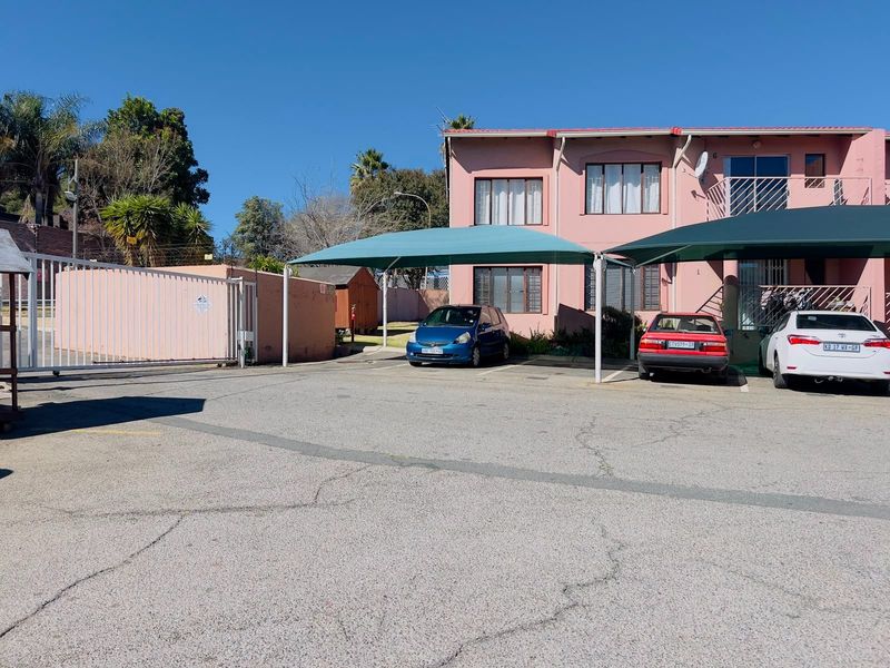 Front exterior view of the townhouse complex featuring a paved driveway, shade cloth carports, and perimeter security fencing.