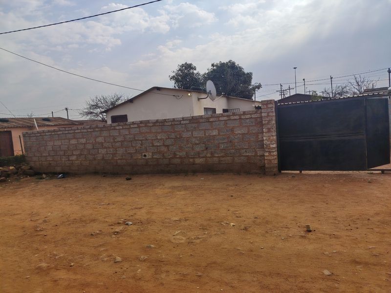 Front view of the property featuring a perimeter brick wall, metal driveway gate, and an unpaved entrance area.