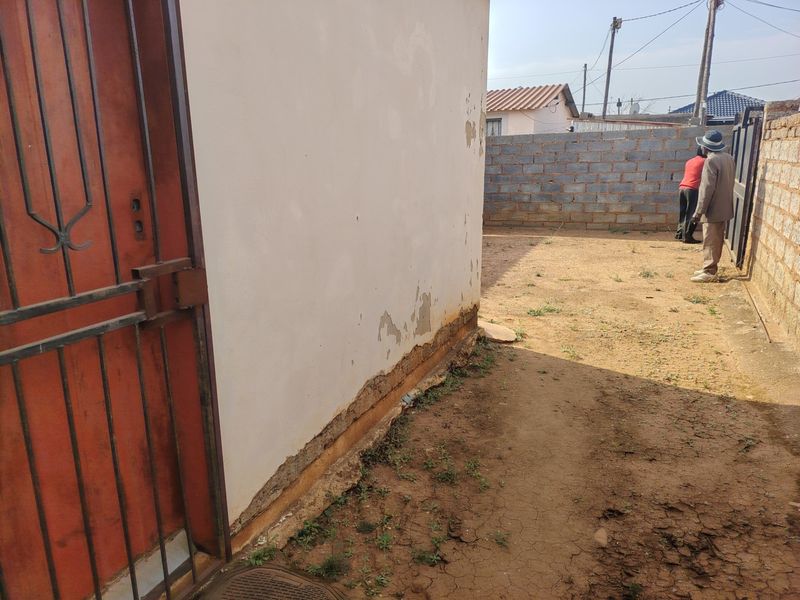Rear exterior view showing a side wall with visible paint peeling and foundation deterioration, alongside a boundary wall and metal gate.