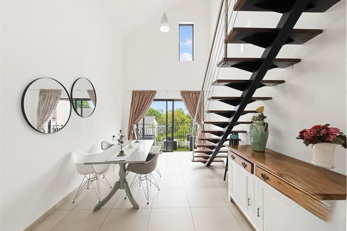 Bright open-plan living area featuring a striking open-tread staircase, high vaulted ceilings, tiled flooring, and sliding doors leading to an outdoor balcony.