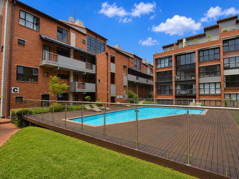A well-maintained communal pool area featuring timber decking, glass balustrades, and views of the surrounding brick apartment complex.