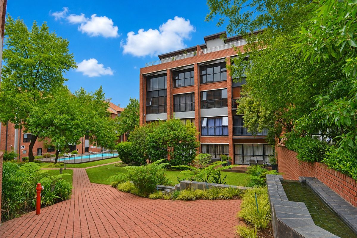 A view of the apartment complex exterior featuring a brick facade, landscaped gardens, paved walkways, a water feature, and a communal swimming pool.
