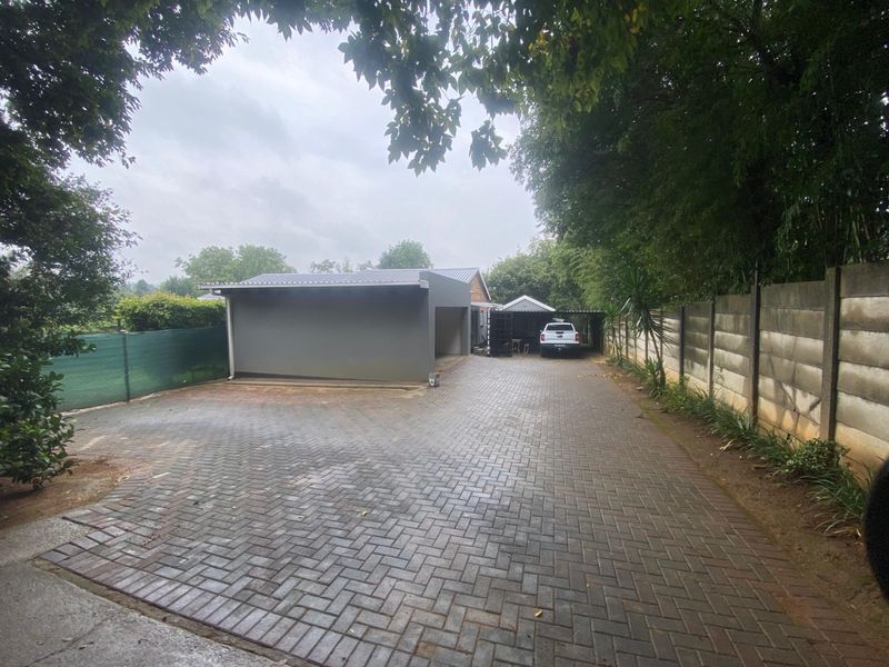 A spacious paved driveway leading to a carport and garage area, enclosed by a concrete boundary wall and mature trees.