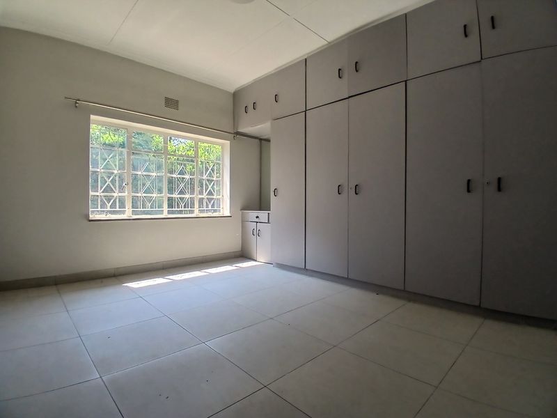 A bright, empty bedroom featuring neutral tiled flooring and extensive built-in cupboard storage with security bars on the window.