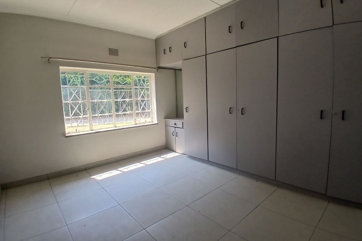 A bright, empty bedroom featuring neutral tiled flooring and extensive built-in cupboard storage with security bars on the window.