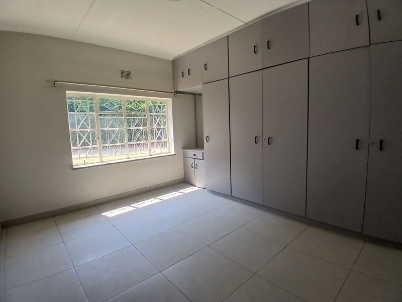 A bright, empty bedroom featuring neutral tiled flooring and extensive built-in cupboard storage with security bars on the window.