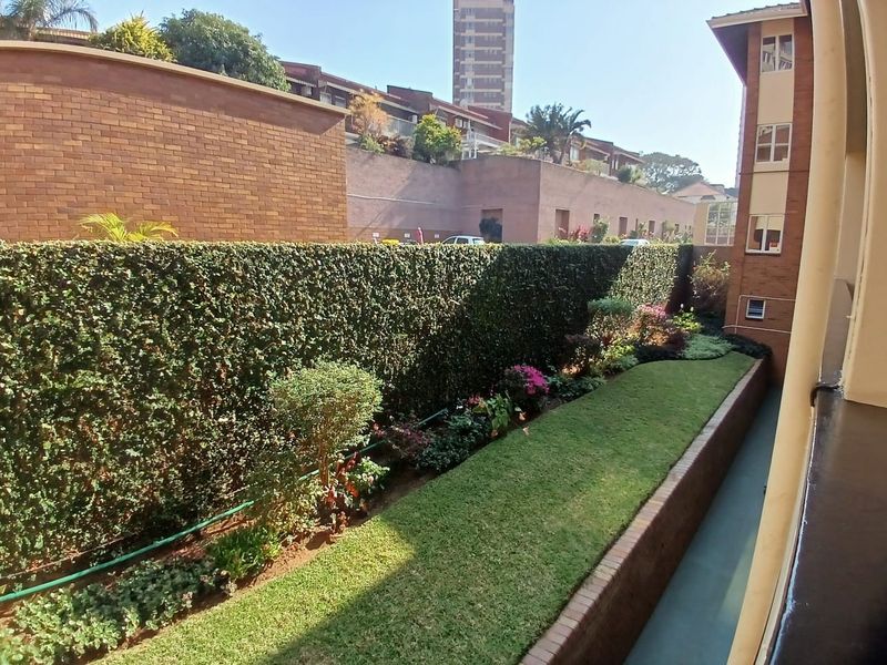 A well-maintained garden area featuring a manicured lawn, established hedge, and brick retaining wall, viewed from an elevated position.