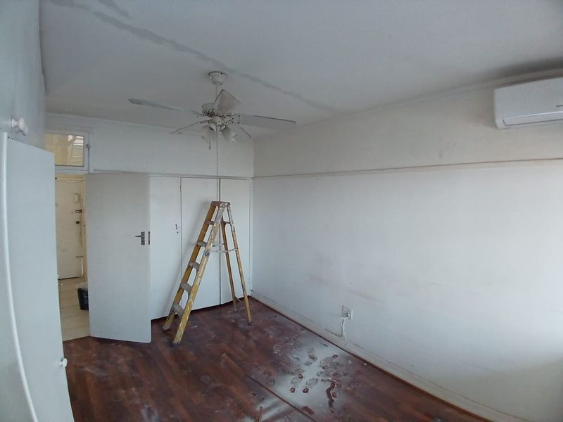 An empty bedroom featuring built-in cupboards, a ceiling fan, and a wall-mounted air conditioning unit, currently requiring cosmetic attention to walls and flooring.