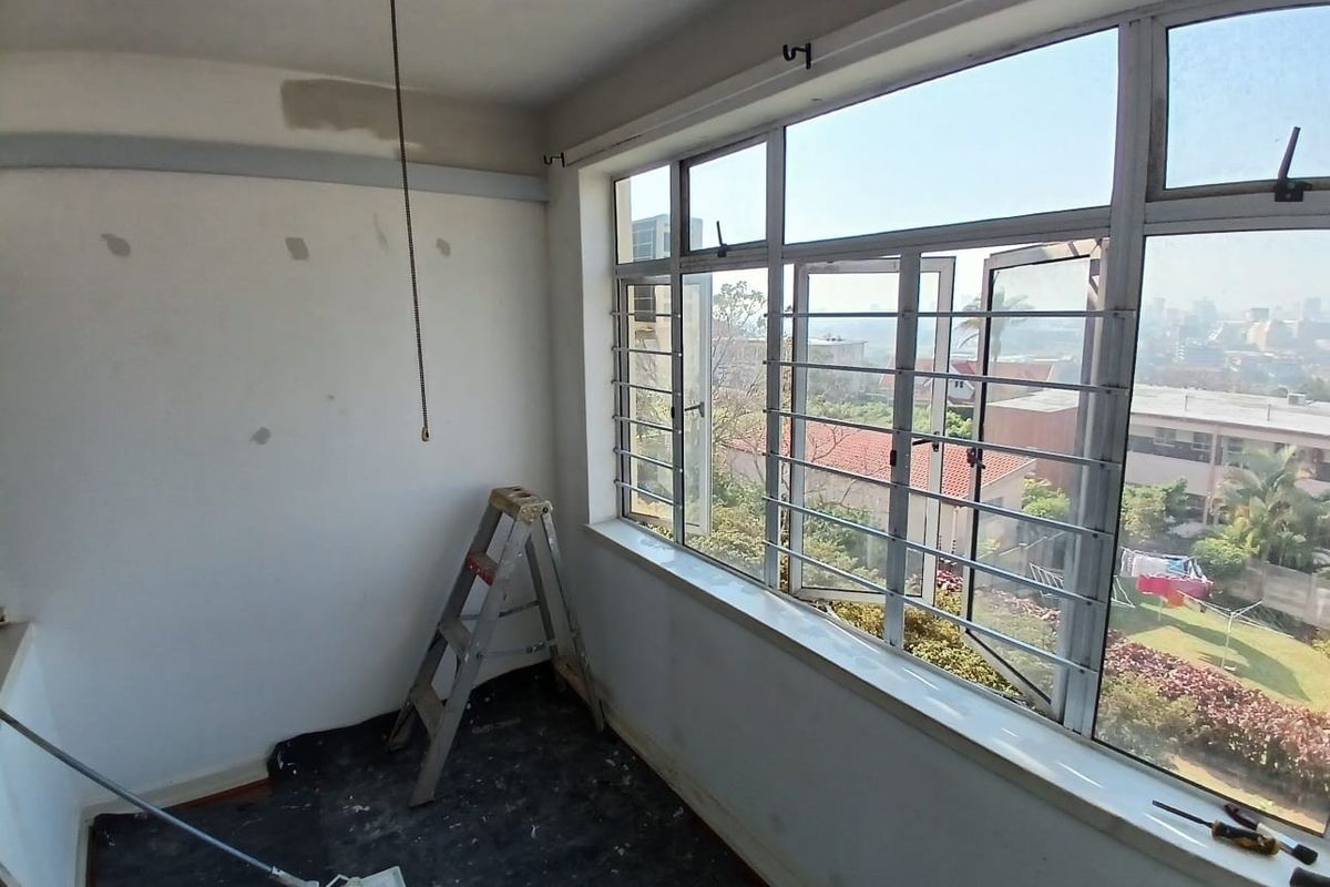 An empty bedroom featuring large aluminium windows with security bars, a ceiling fan, and walls currently undergoing preparation for painting.