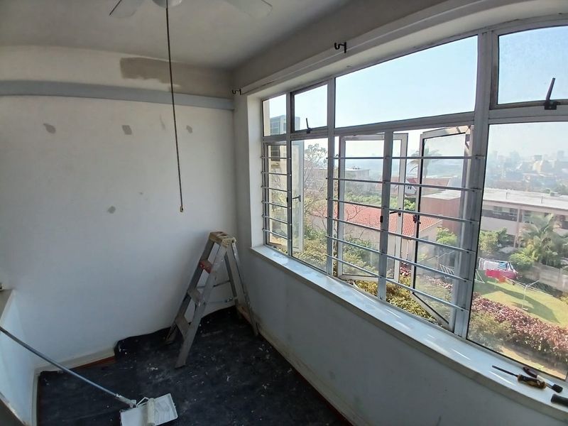 An empty bedroom featuring large aluminium windows with security bars, a ceiling fan, and walls currently undergoing preparation for painting.