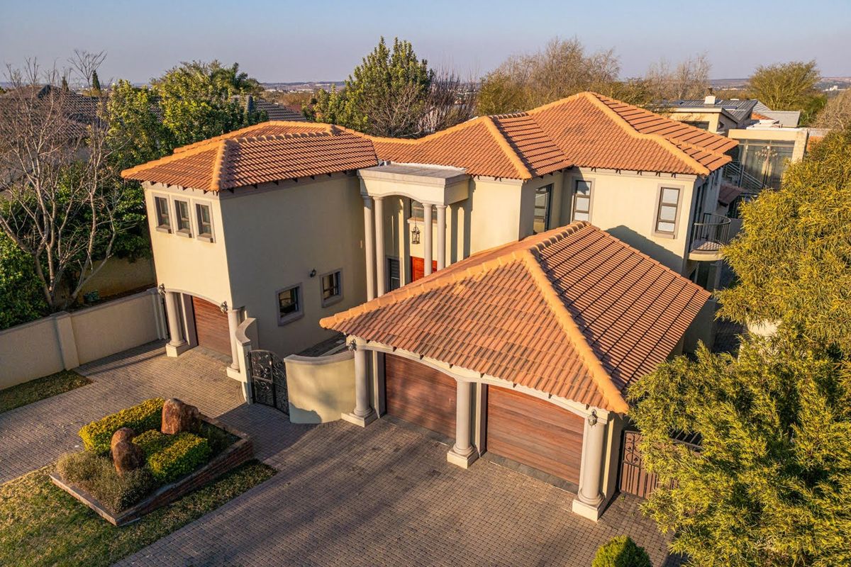 Aerial view of a multi-level residential property featuring a terracotta tile roof, double garage, paved driveway, and classic architectural columns.