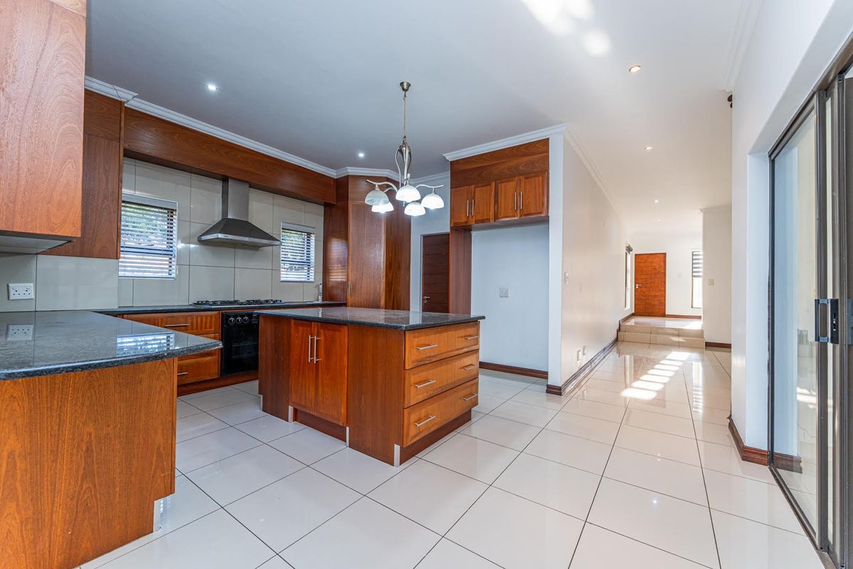 A spacious, modern kitchen featuring warm wooden cabinetry, granite countertops, a central island, and sleek tiled flooring, leading into an open-plan living area.