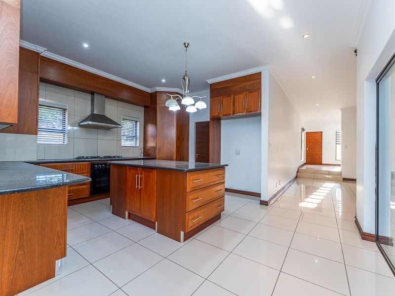 A spacious, modern kitchen featuring warm wooden cabinetry, granite countertops, a central island, and sleek tiled flooring, leading into an open-plan living area.