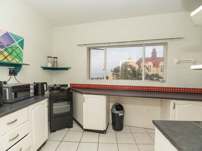 A functional kitchen featuring white cabinetry, dark laminate countertops, a red tiled backsplash, and tiled flooring with a view through the sliding window.