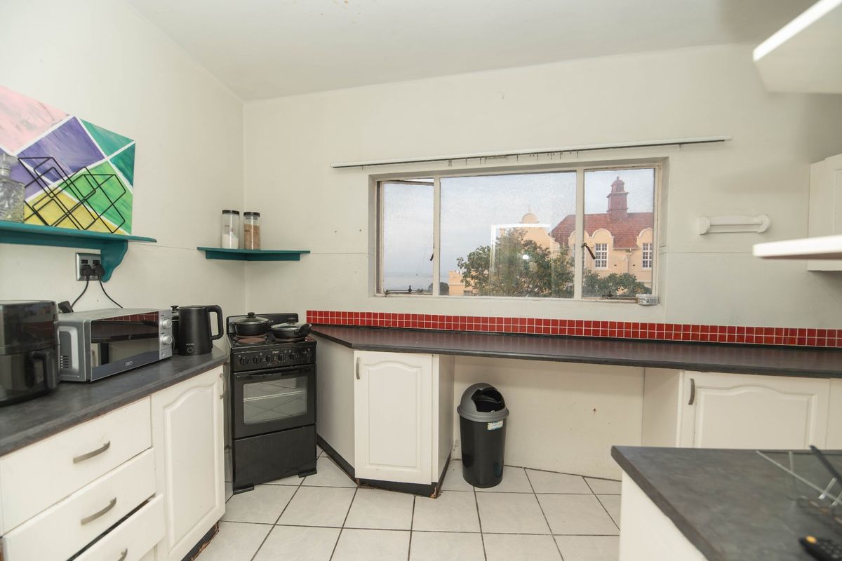 A functional kitchen featuring white cabinetry, dark laminate countertops, a red tiled backsplash, and tiled flooring with a view through the sliding window.