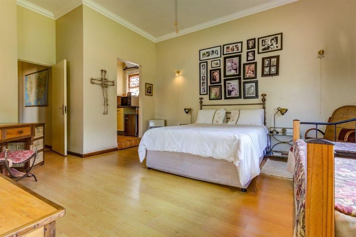 A spacious bedroom featuring laminate flooring, coving, and wall sconces, with an adjoining view into a secondary room or kitchen area.