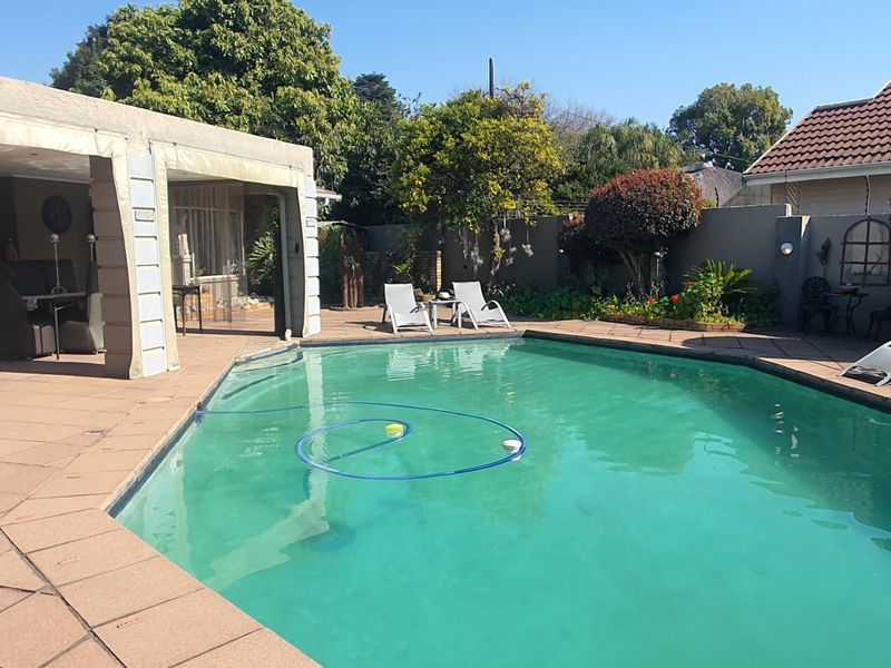 A well-maintained outdoor pool area featuring a paved deck, a covered patio space, and surrounding boundary walls with established garden landscaping.