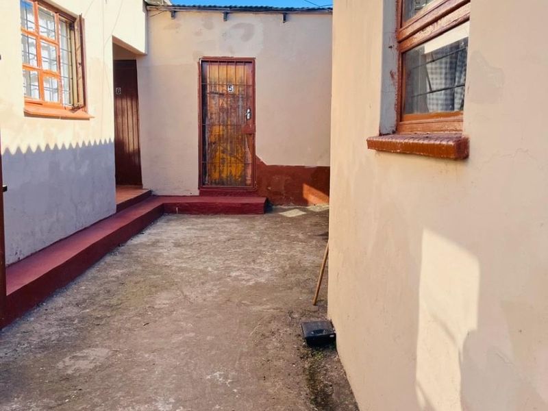 A narrow rear courtyard area featuring concrete paving, weathered exterior walls, and security-gated access points between building structures.