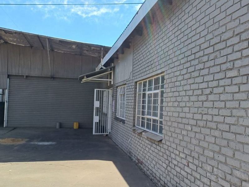 Exterior view of an industrial property featuring brick construction, steel security gates, metal-framed windows, and a large roller shutter door access point.