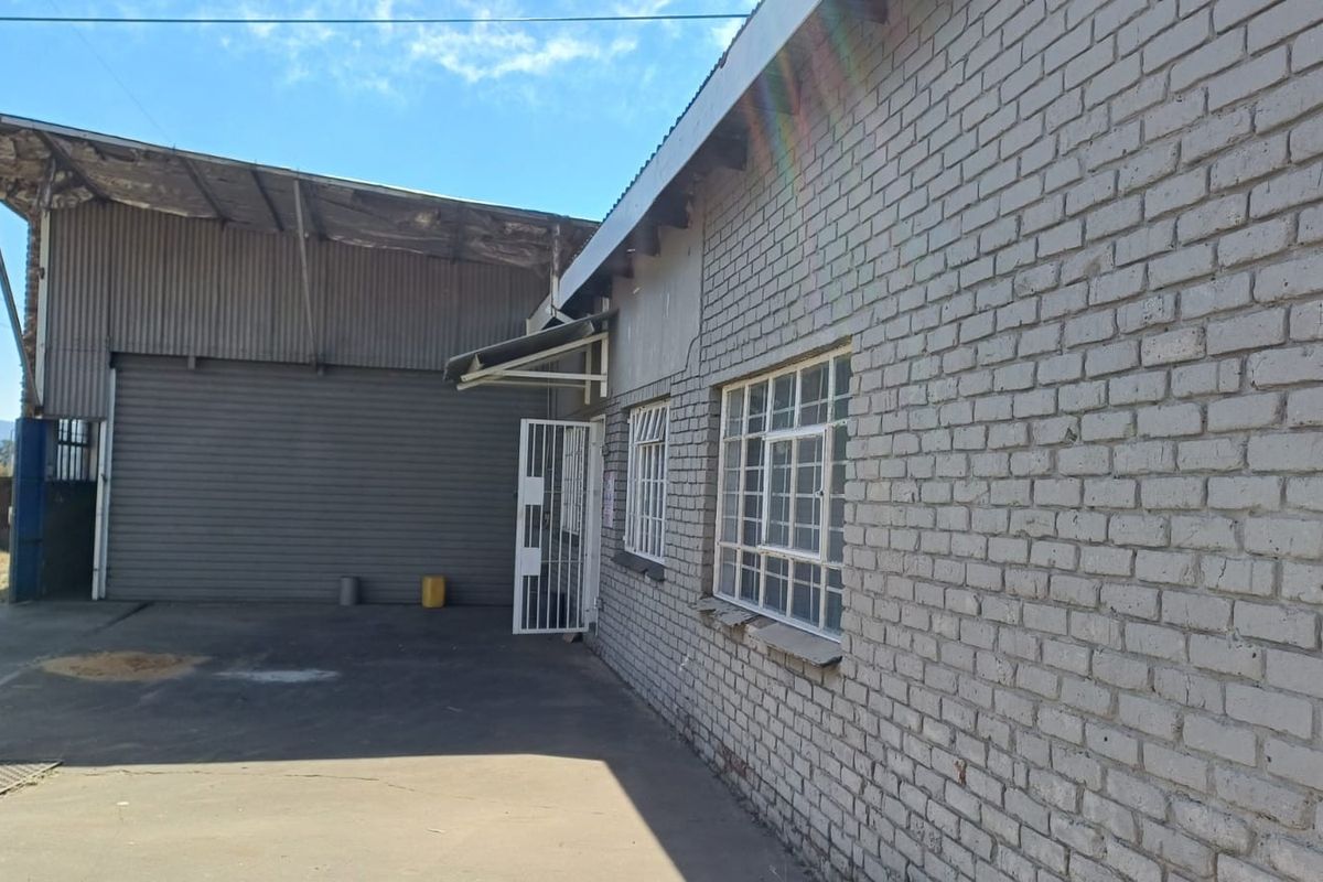 Exterior view of an industrial property featuring brick construction, steel security gates, metal-framed windows, and a large roller shutter door access point.