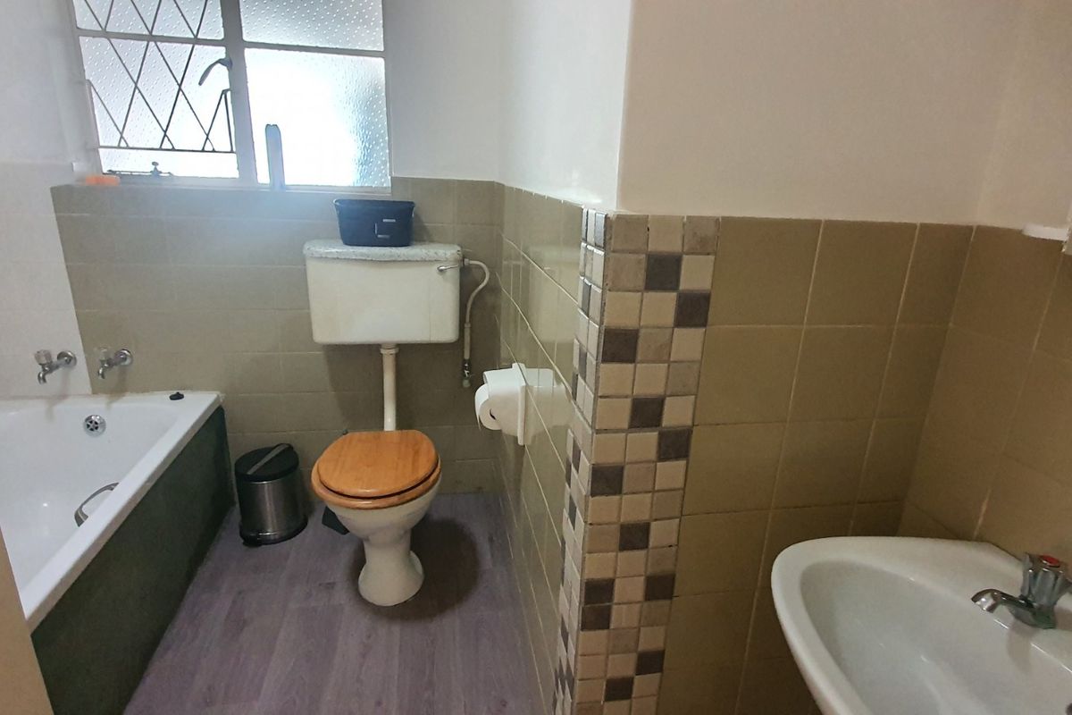 A functional bathroom featuring a bathtub, pedestal basin, and toilet with dated wall tiling and security bars on the window.