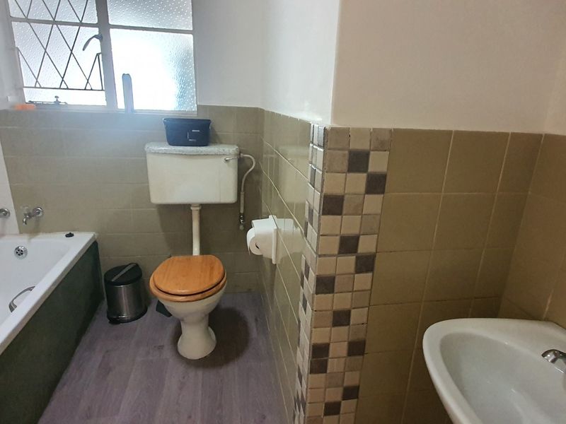 A functional bathroom featuring a bathtub, pedestal basin, and toilet with dated wall tiling and security bars on the window.