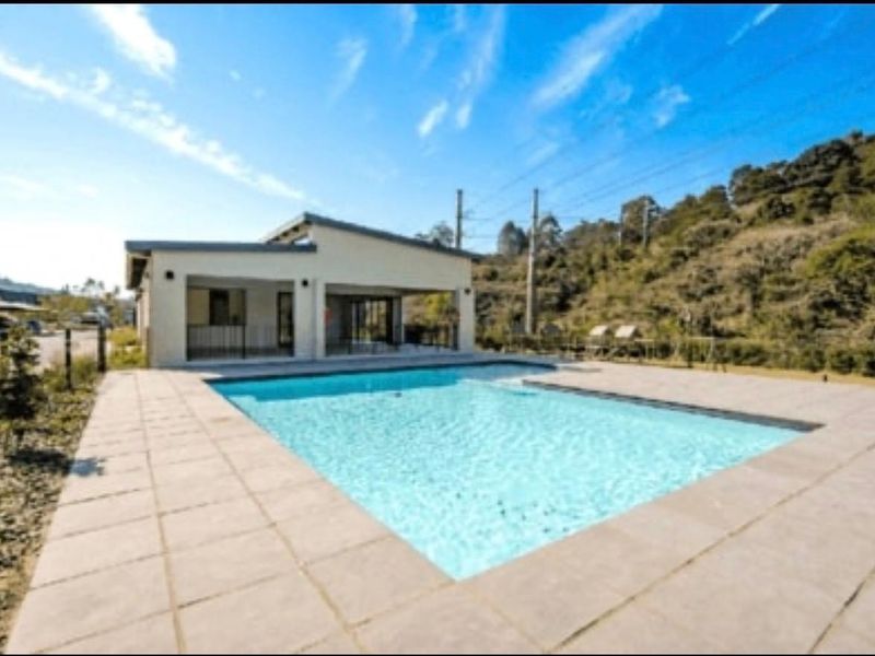 A well-maintained outdoor swimming pool area featuring a paved deck and a covered patio space, set against a natural hillside backdrop.