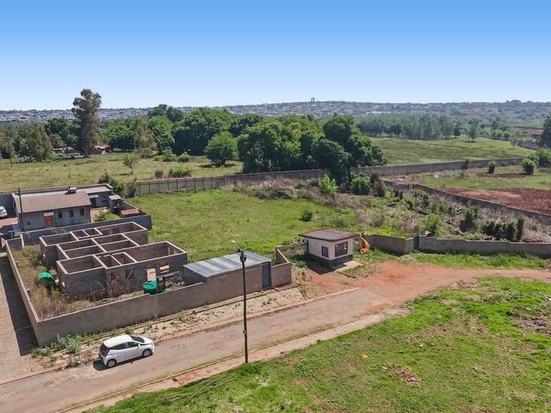 Aerial view of a commercial property featuring a perimeter wall, security gate, paved driveway, a small guard house, and an uncompleted masonry building structure.